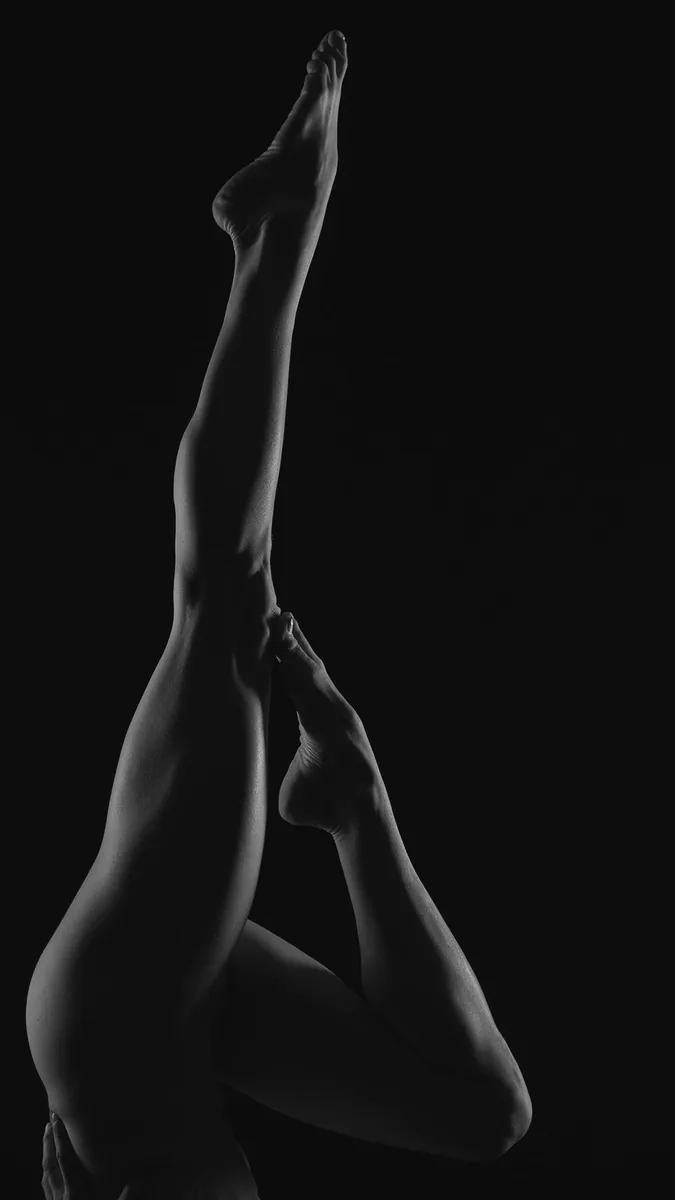 close-up of a personâ€™s legs in a vertical pose with one leg extended straight up and the other bent, set against a black background with soft lighting highlighting muscle contours