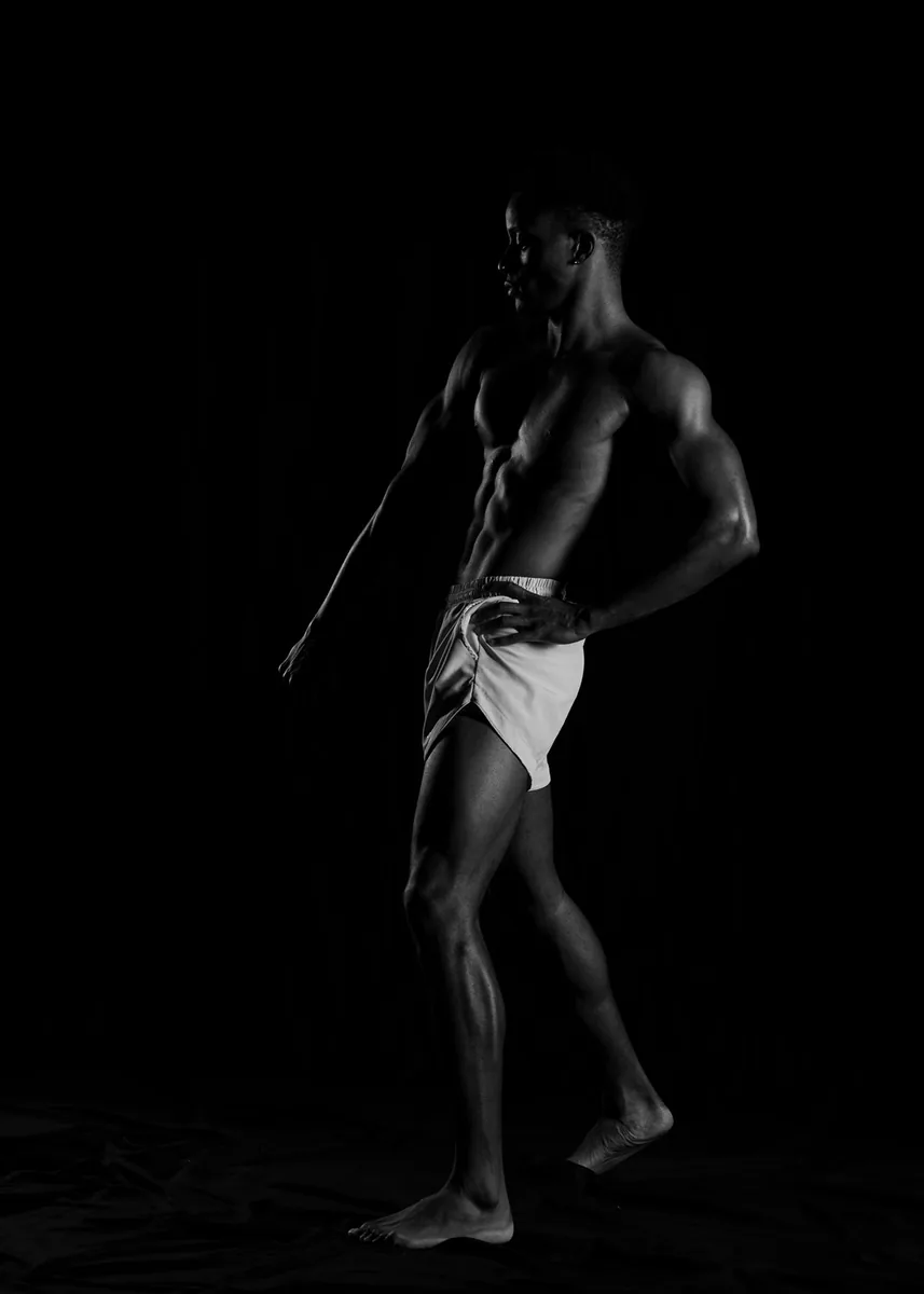 Athletic person standing barefoot, wearing light shorts, showcasing muscular build and defined abs against a dark background