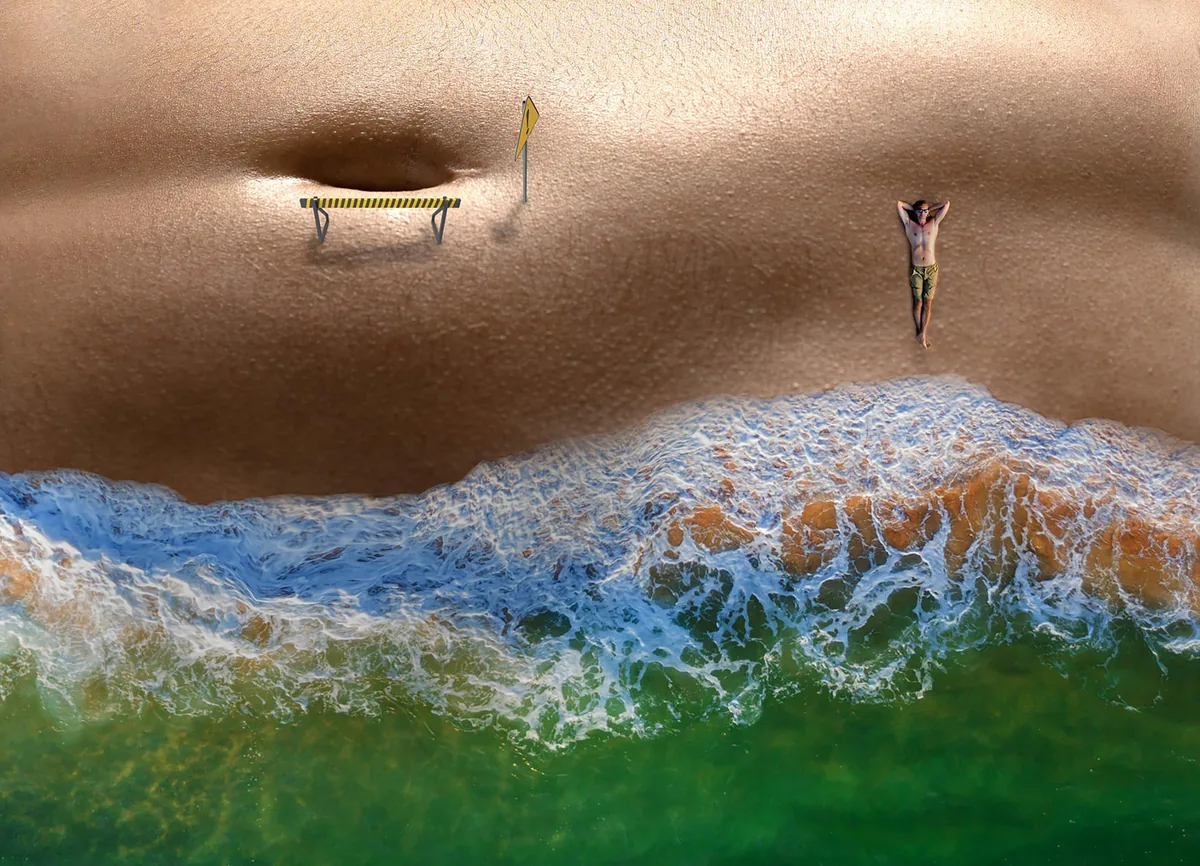 Top-down view of a person lying on sandy beach near foamy ocean waves with a caution barrier and sign on the sand nearby
