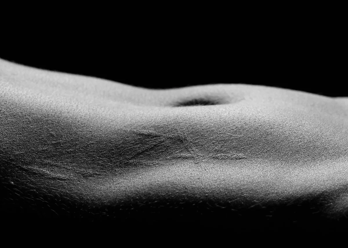 Close-up view of human torso skin showing fine texture, stretch marks, and navel, with dramatic lighting highlighting curves against a dark background
