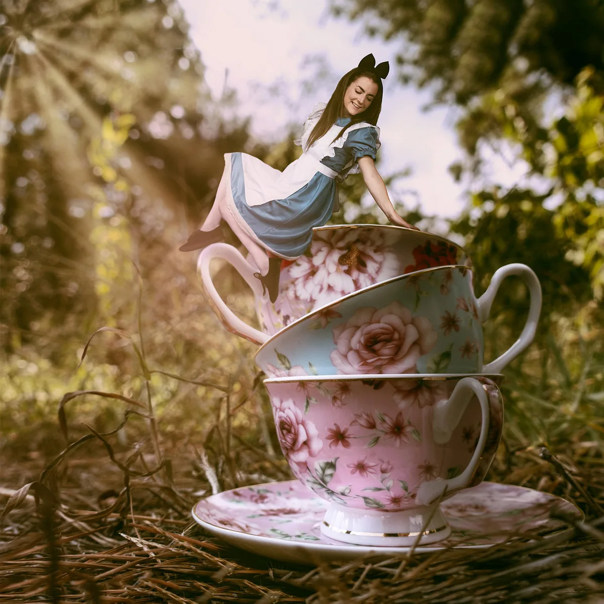 Woman dressed as Alice from Alice in Wonderland sitting on the rim of large floral teacups stacked outdoors amidst grass and pine straw