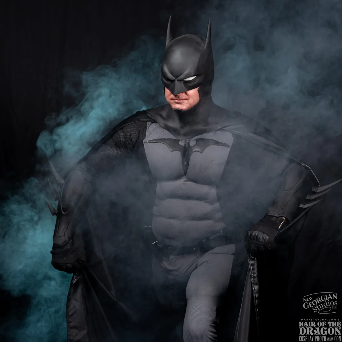 Person dressed as Batman in a detailed costume with cape spreading, surrounded by blue-gray smoke on a dark background