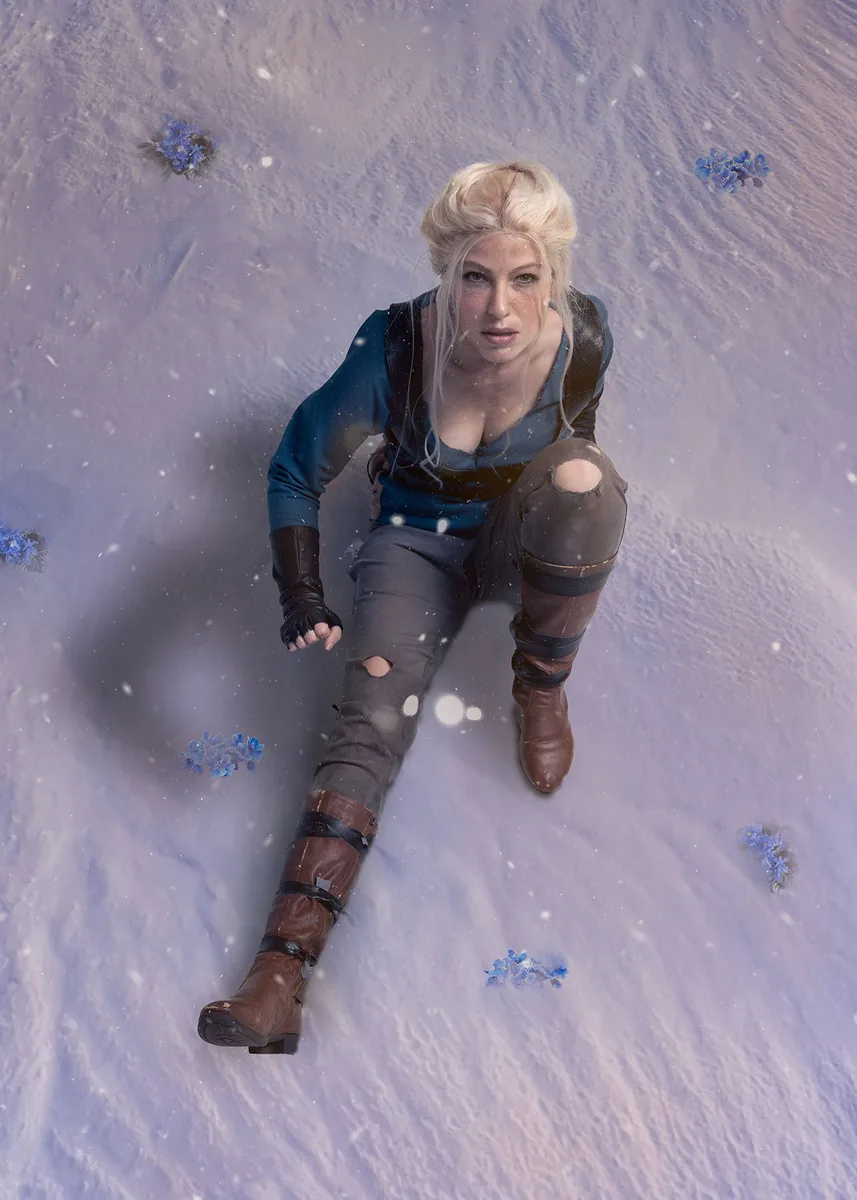 A blonde woman with braided hair kneels on snow-covered ground, wearing a blue shirt, torn gray pants, brown boots, and black fingerless gloves, surrounded by scattered blue flowers and softly falling snow.