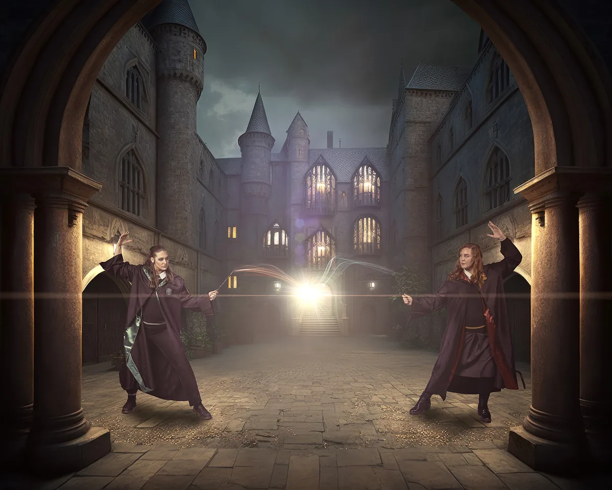 Two women in long dark robes casting magic with wands, facing each other in a stone courtyard of a castle-like building under a cloudy sky, magic light glowing between their wands