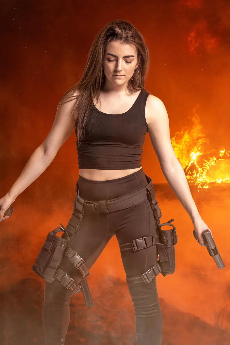 Confident woman with long hair dressed in black tactical gear holding two guns with flames and smoke in the background