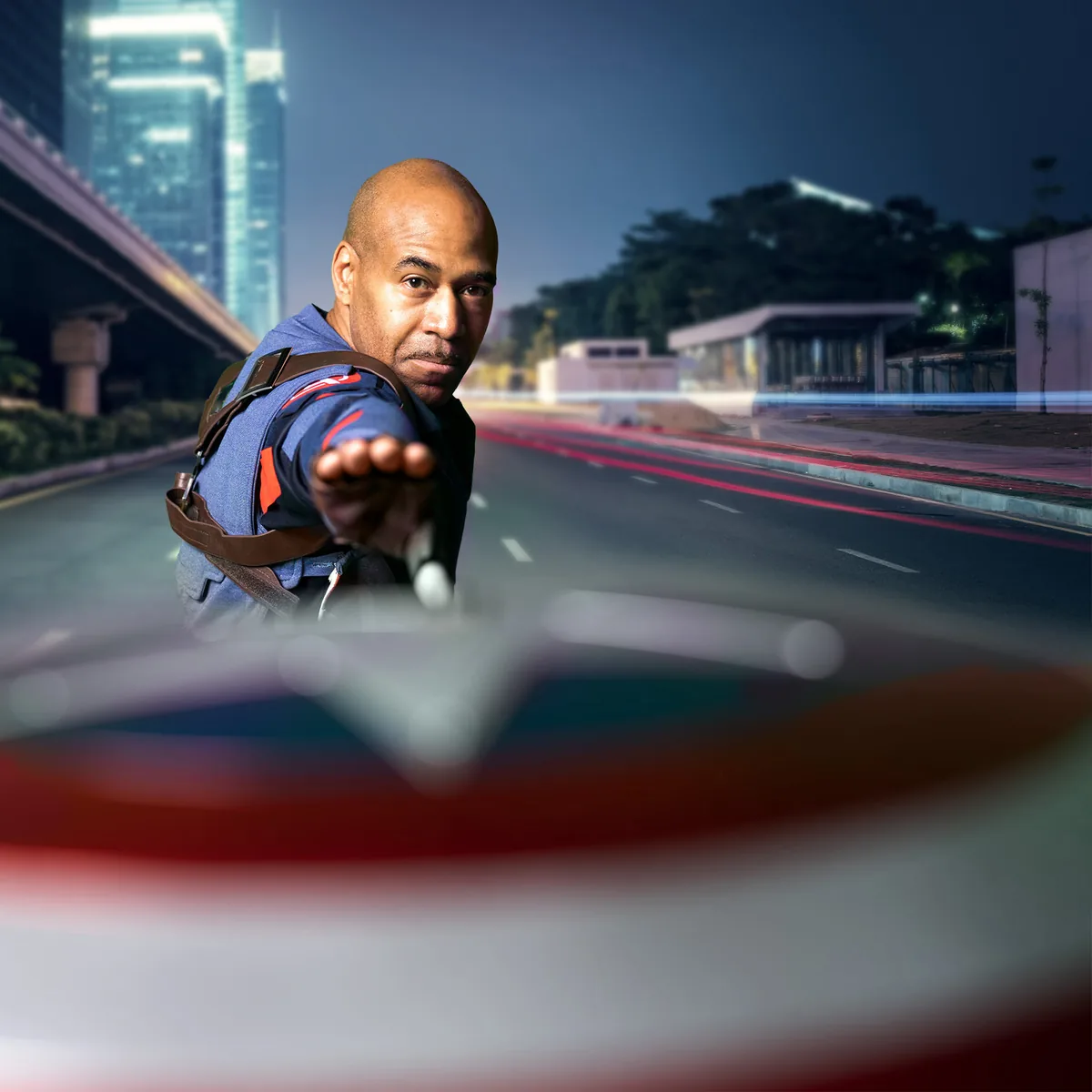 Man in superhero outfit with blue shirt and brown harness reaching forward, with a blurred Captain America-style shield in the foreground, set on a deserted city street at night with buildings and light trails in the background