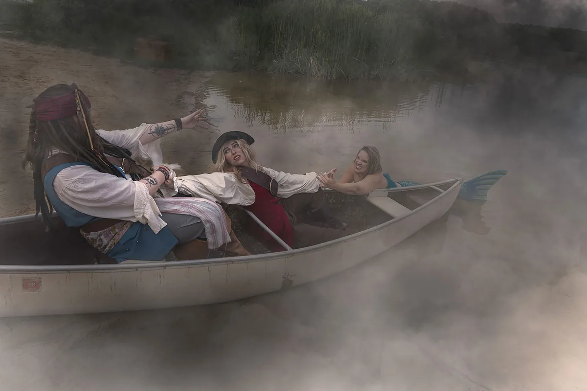 Two people dressed as pirates struggle in a canoe while a woman dressed as a mermaid pulls one pirate's hand from the water, surrounded by thick mist on a lake shore