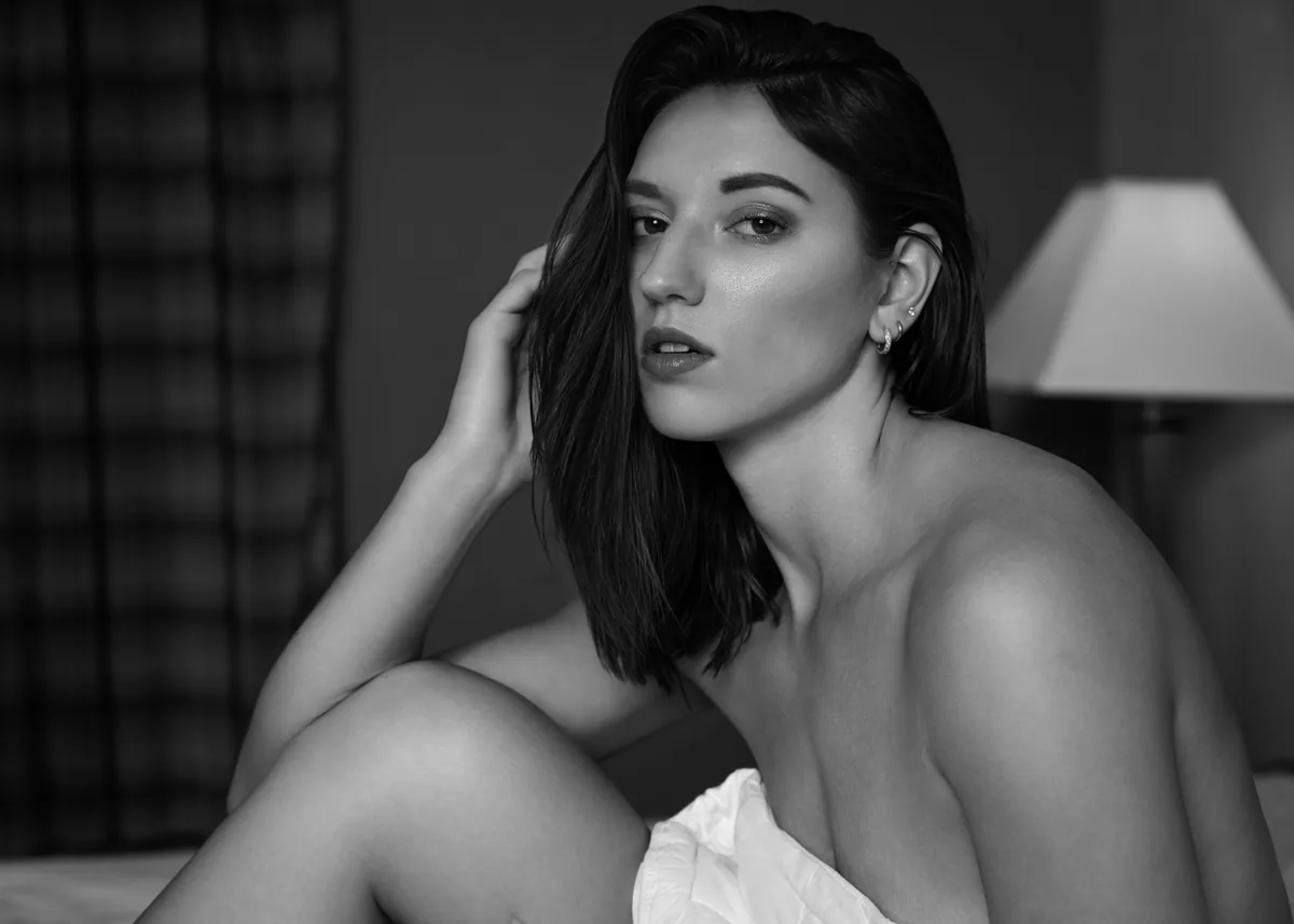 A black and white portrait of a woman sitting with one arm resting on her knee, her head slightly tilted, with soft lighting highlighting her face and bare shoulder, indoors near a lamp.