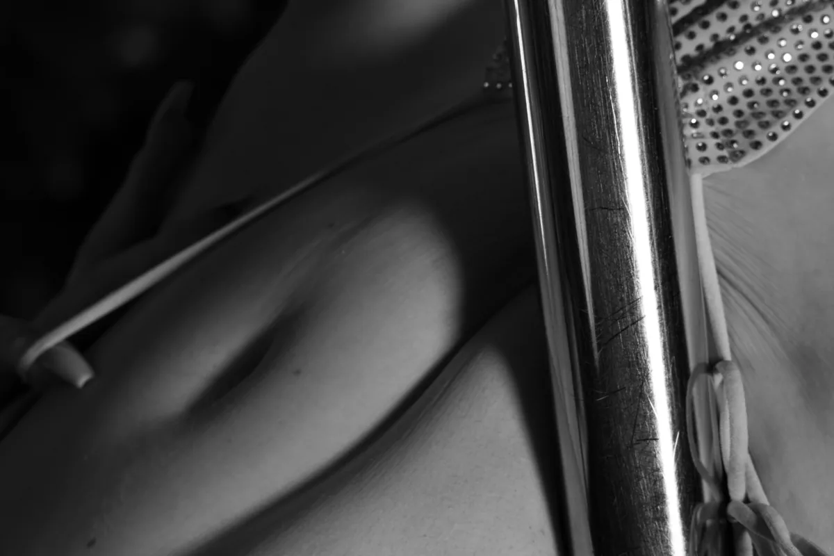 Black and white close-up of a shoulder and upper chest partially covered by a strap with a reflective metal pole positioned vertically in the foreground, highlighting textures and shadows.