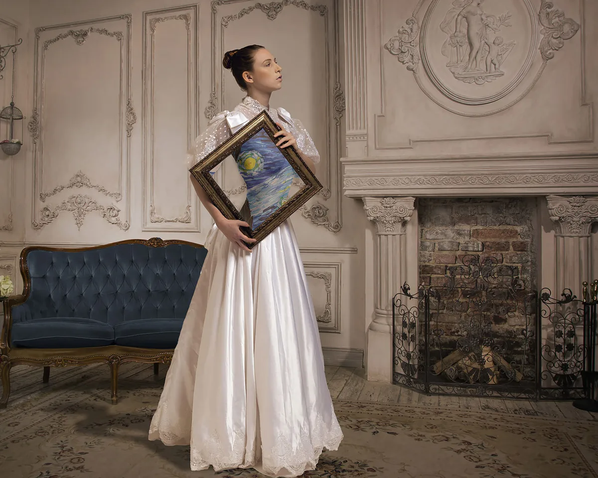 Woman in a white vintage dress holding a diamond-shaped frame displaying a Van Gogh style painting, standing in a room with ornate wall paneling, a blue velvet sofa, and a fireplace with decorative molding