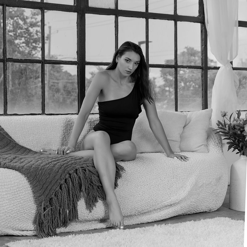Woman with long hair wearing a one-shoulder black swimsuit sitting on a textured sofa with a throw blanket, large window with multiple panes behind her, and a tall vase with flowers to the side