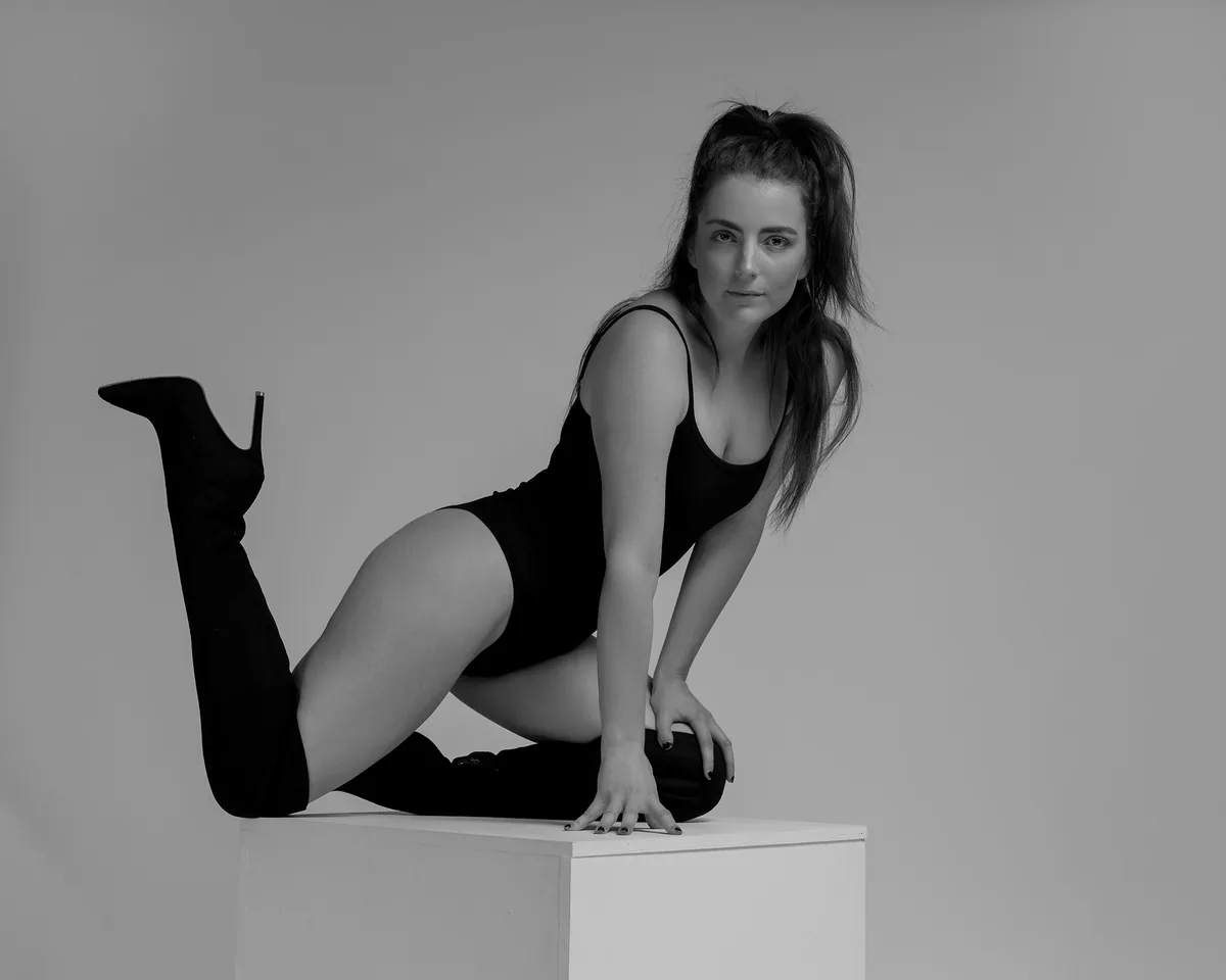 Woman kneeling on a white cube wearing a black bodysuit and thigh-high black boots, with one leg lifted, against a plain gray background