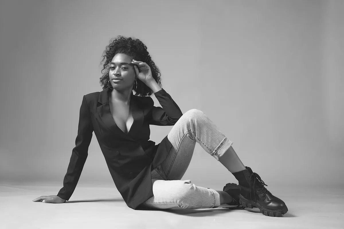 A woman with curly hair sits on the floor against a plain background. She is wearing a dark blazer, light-colored rolled-up jeans, and chunky black boots. One hand rests on the floor while her other hand lightly touches her forehead, exuding confidence and calm.