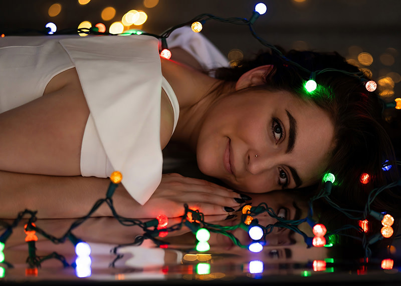 A woman lies on a reflective surface with her head resting on her folded hands. She wears a white garment and has a small nose stud. Colorful string lights in red, green, blue, and orange are draped around her head and reflected on the surface, creating a festive and calm atmosphere.