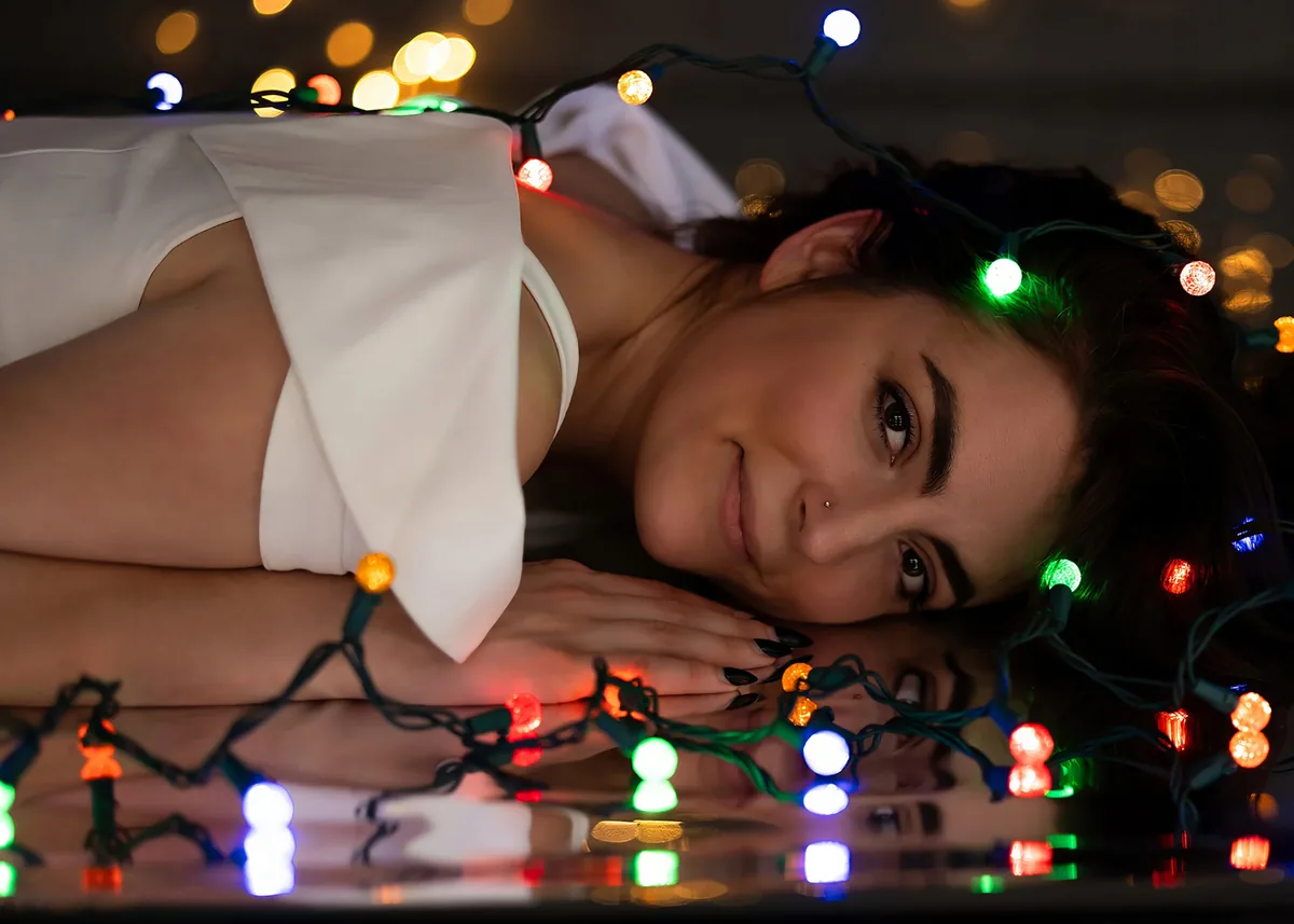 A woman lies on a reflective surface with her head resting on her folded hands. She wears a white garment and has a small nose stud. Colorful string lights in red, green, blue, and orange are draped around her head and reflected on the surface, creating a festive and calm atmosphere.