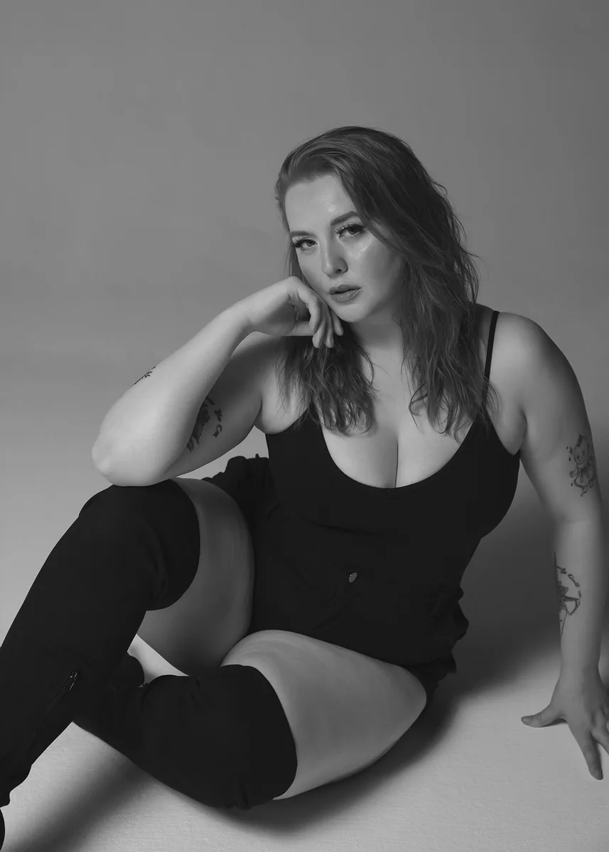 Woman with shoulder-length hair seated on the floor wearing a black sleeveless top, black thigh-high boots, and shorts, resting her head on her hand with a thoughtful expression, tattoos visible on both arms, against a plain backdrop