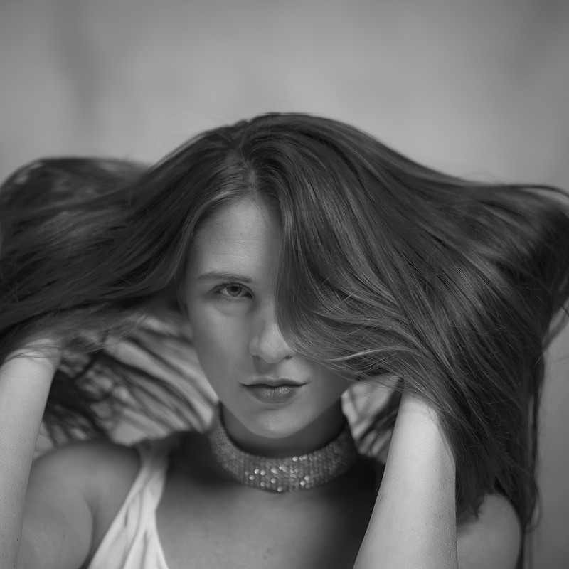 Woman with long hair lifting and flipping it with both hands, wearing a sleeveless top and a sparkling choker necklace, looking directly ahead