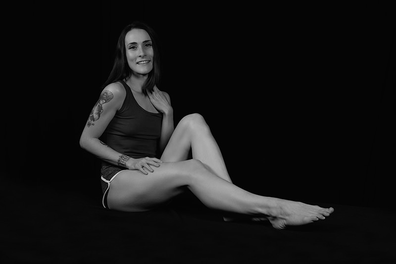 Smiling woman with dark hair sitting on floor against black background, wearing tank top and shorts, showing tattoos on left arm and wrist