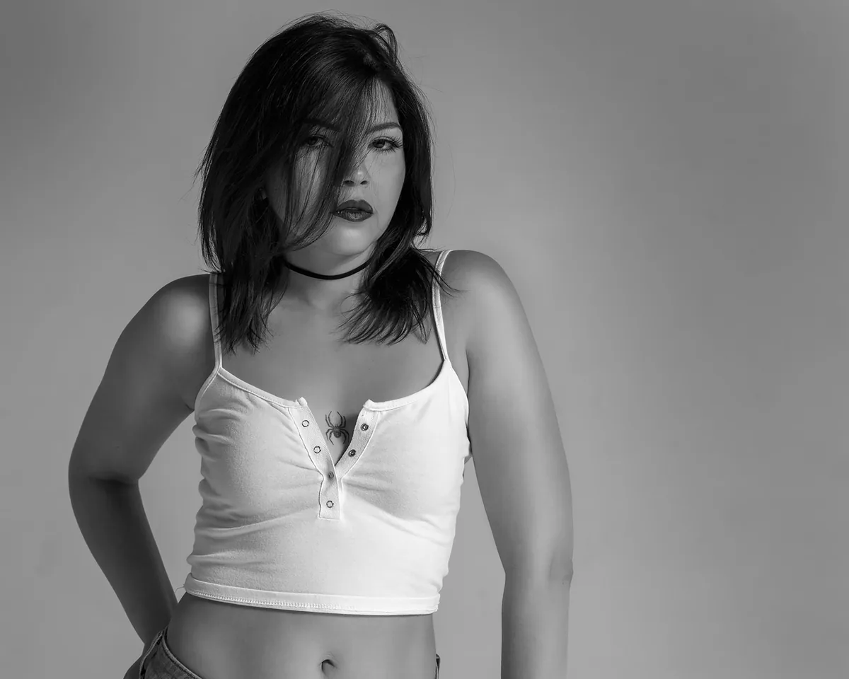 Young woman with medium-length dark hair partially covering her face, wearing a white crop top with a spider tattoo on her chest, and a black choker, standing against a plain background