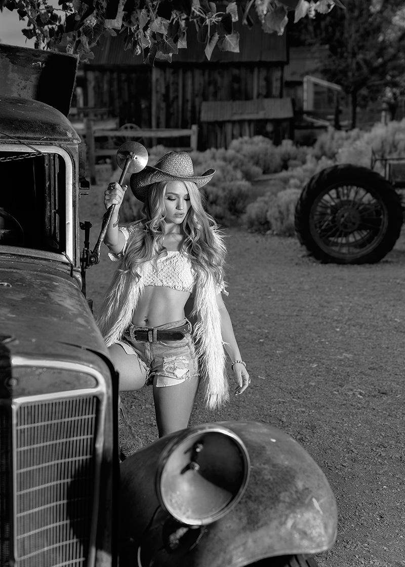 Woman wearing a cowboy hat, cropped lace top, fur vest, and denim shorts leaning against an old vintage car, holding a car horn, in a rustic outdoor setting with bushes and an old wooden building in the background