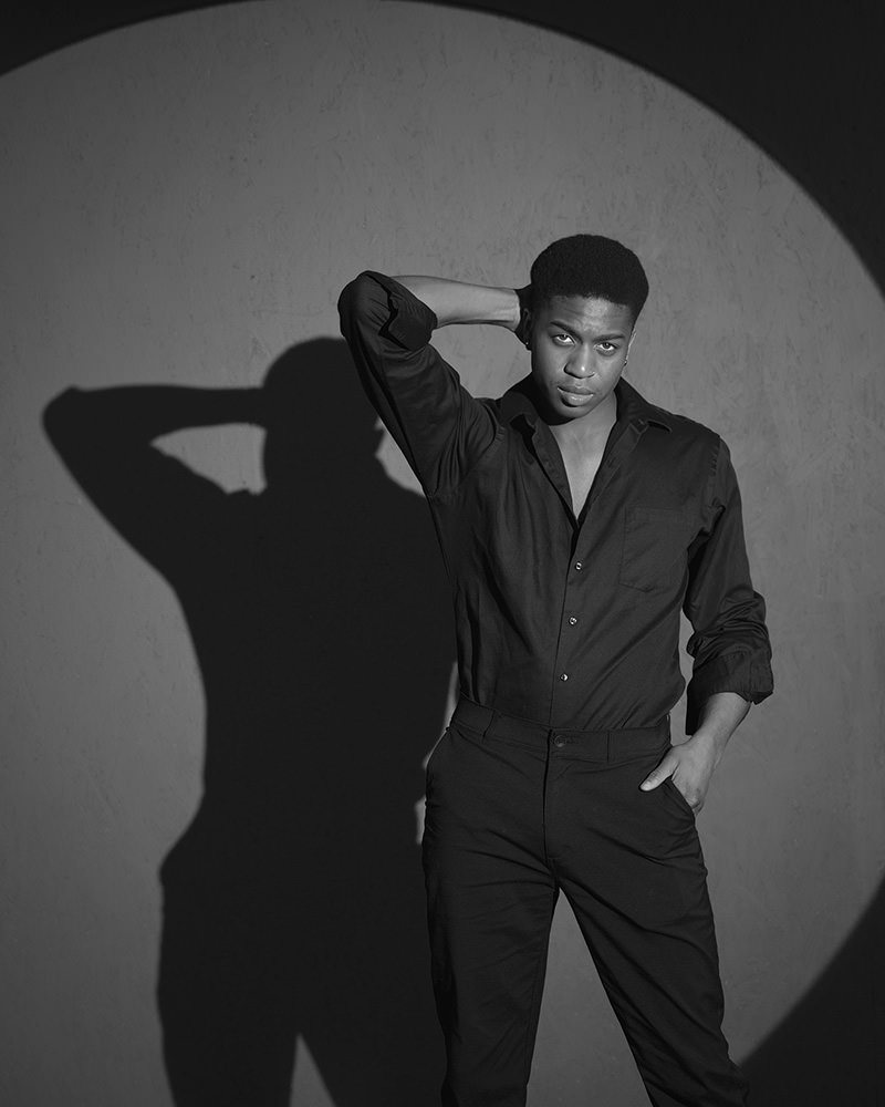 Young man wearing a black shirt and pants standing with one hand behind his neck and the other in his pocket, illuminated by a circular spotlight casting a shadow on the textured wall behind him
