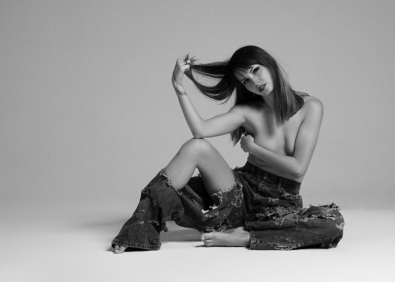 Young woman sitting on the floor with torn denim pants, topless, holding her hair with one hand and covering her chest with the other, against a plain backdrop