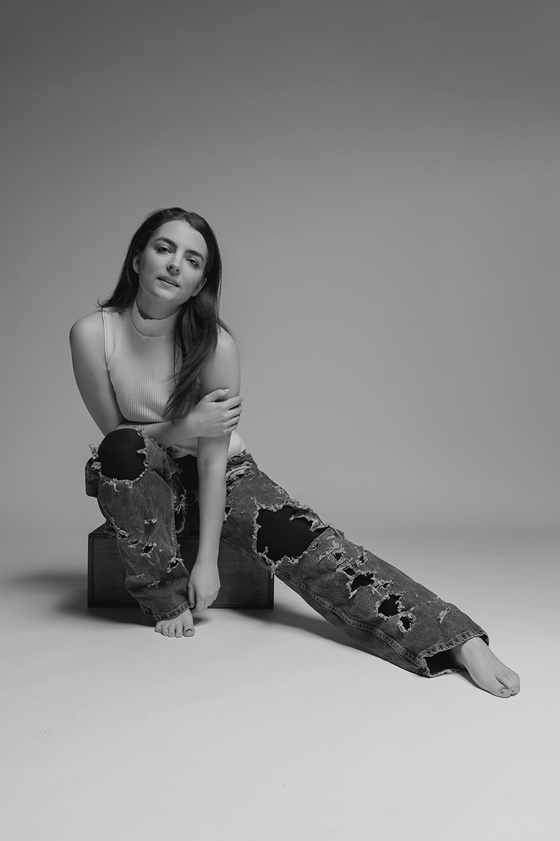 Young woman sitting on a low wooden box against a plain background, wearing a sleeveless ribbed top and heavily ripped jeans, barefoot with one leg bent and the other stretched out, looking directly at the camera with a slight smile