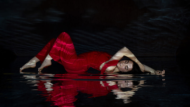 Woman lying on her side in a red dress with water ripples reflecting across her body and the surface beneath her in a dark setting