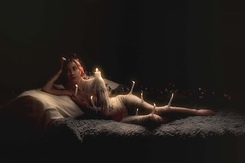 Person reclining on a soft surface, covered in dripping wax, with lit candles affixed to their body, surrounded by a dark background with scattered flower petals