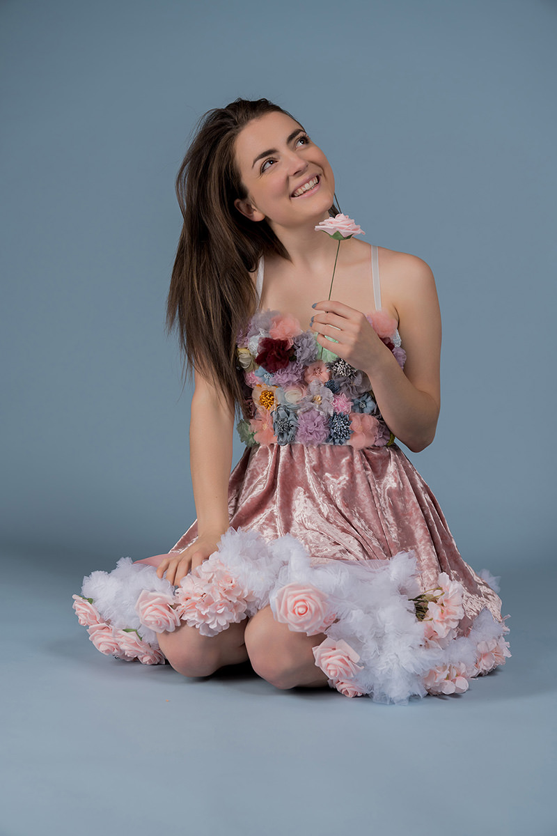 Smiling woman with long brown hair kneeling on the floor, wearing a pink velvet dress adorned with multicolored fabric flowers on the bodice and white tulle with pink roses on the skirt, holding a single pink rose near her face and looking upwards against a solid gray-blue background.