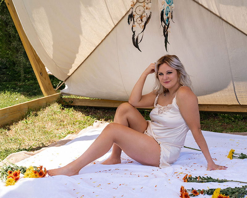 Woman with blonde hair wearing a white satin camisole and shorts sitting on a white blanket outdoors near a cream-colored tent, surrounded by scattered orange and yellow flowers and hanging dreamcatchers above her head