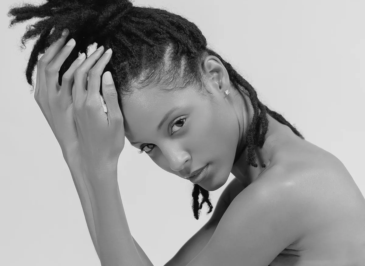 Close-up of a woman with dreadlocks tied up, looking directly at the camera with her hands gently holding her hair, bare shoulders visible against a plain background