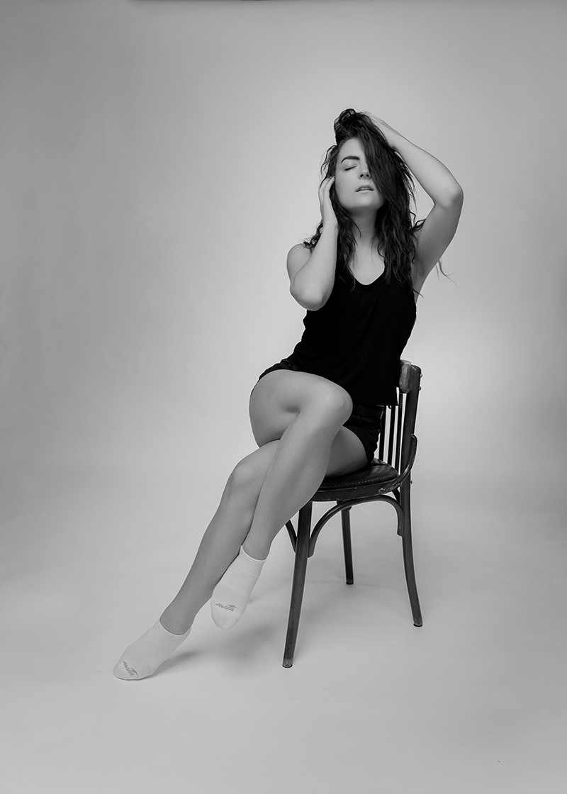 Woman with long, wavy hair sitting on a wooden chair, legs crossed, wearing a black sleeveless top, black shorts, and white socks, with eyes closed and one hand touching her hair.