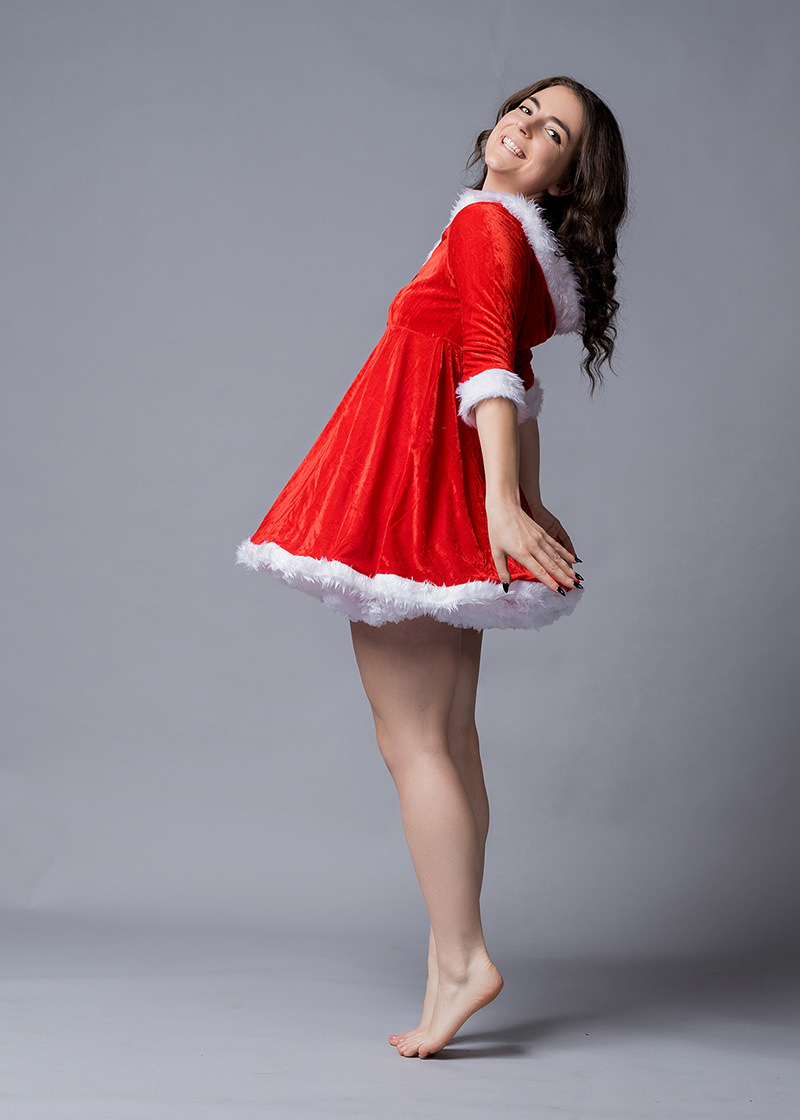 Woman standing on tiptoes, smiling, wearing a red velvet dress with white fur trim and long curly hair against a plain grey background