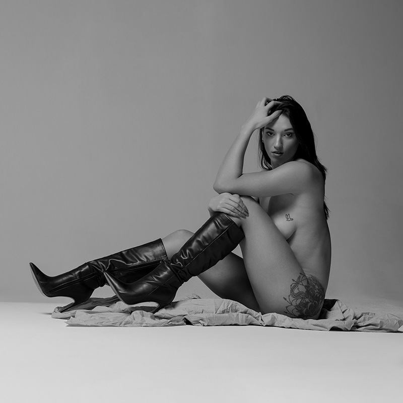 Seated woman wearing tall leather boots with tattoos visible on her side and thigh, against a plain backdrop, posing with one hand on her head and the other resting on her knee