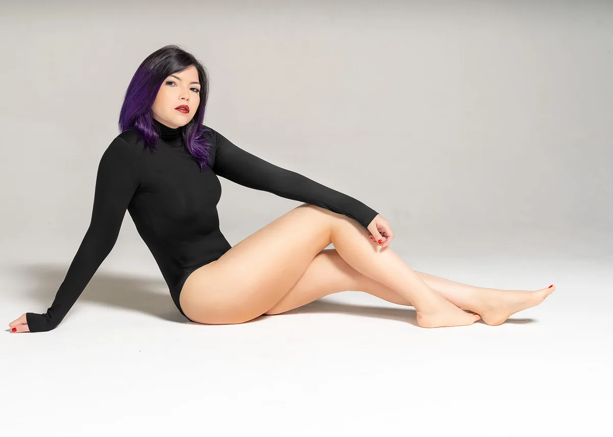 Woman with purple hair sitting on the floor wearing a black long-sleeve bodysuit, posed with one hand behind her and legs extended in front with crossed ankles, against a plain white background