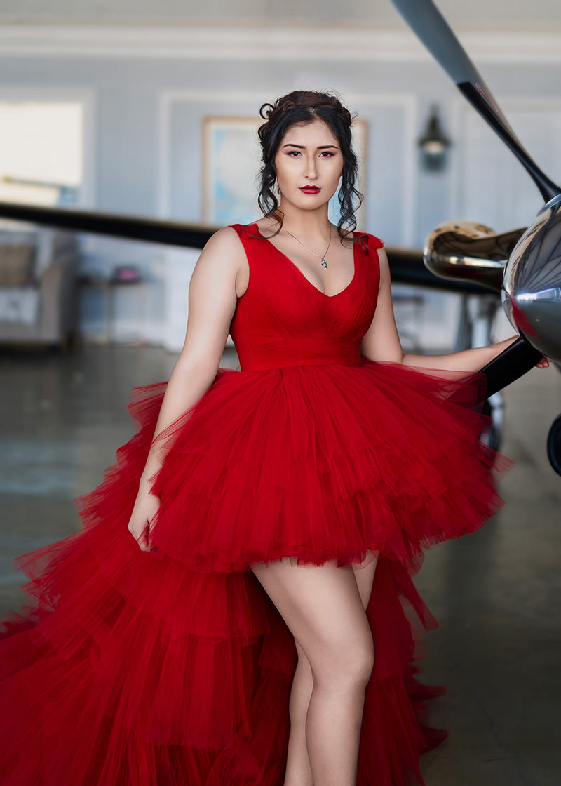 Woman wearing a vibrant red layered tulle gown with a fitted bodice, standing confidently with one hand resting on a shiny airplane propeller in a hangar.