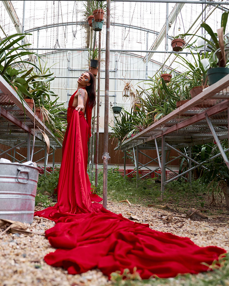 Person in a long flowing red dress standing in a greenhouse between rows of potted plants, holding the fabric of the dress that trails on the ground, with a visible tattoo on the upper arm and a thoughtful expression