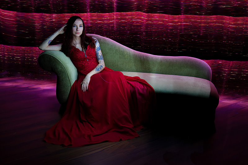 Woman with long dark hair and tattoos reclining on a green velvet chaise lounge, wearing a flowing red dress, with a glowing, wavy red and purple abstract background.