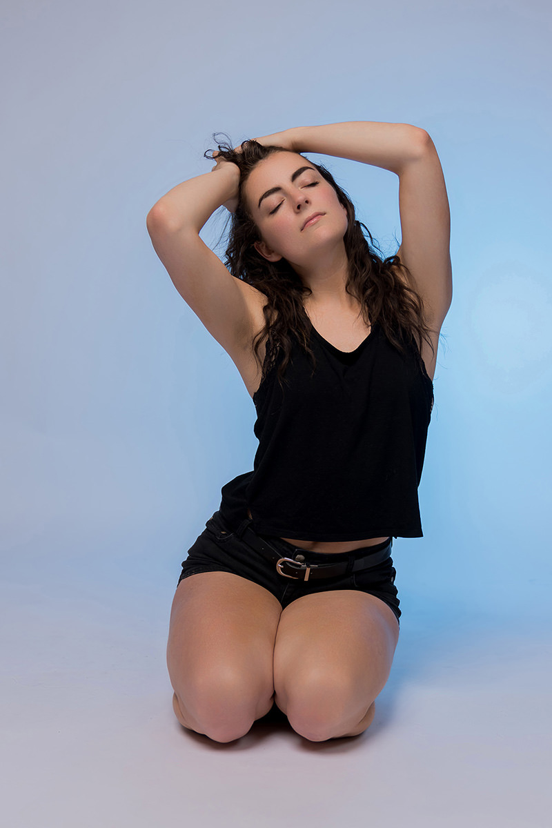 Woman kneeling on the floor with her eyes closed, hands resting on her head, wearing a black sleeveless top and black shorts, with a calm expression against a light blue background