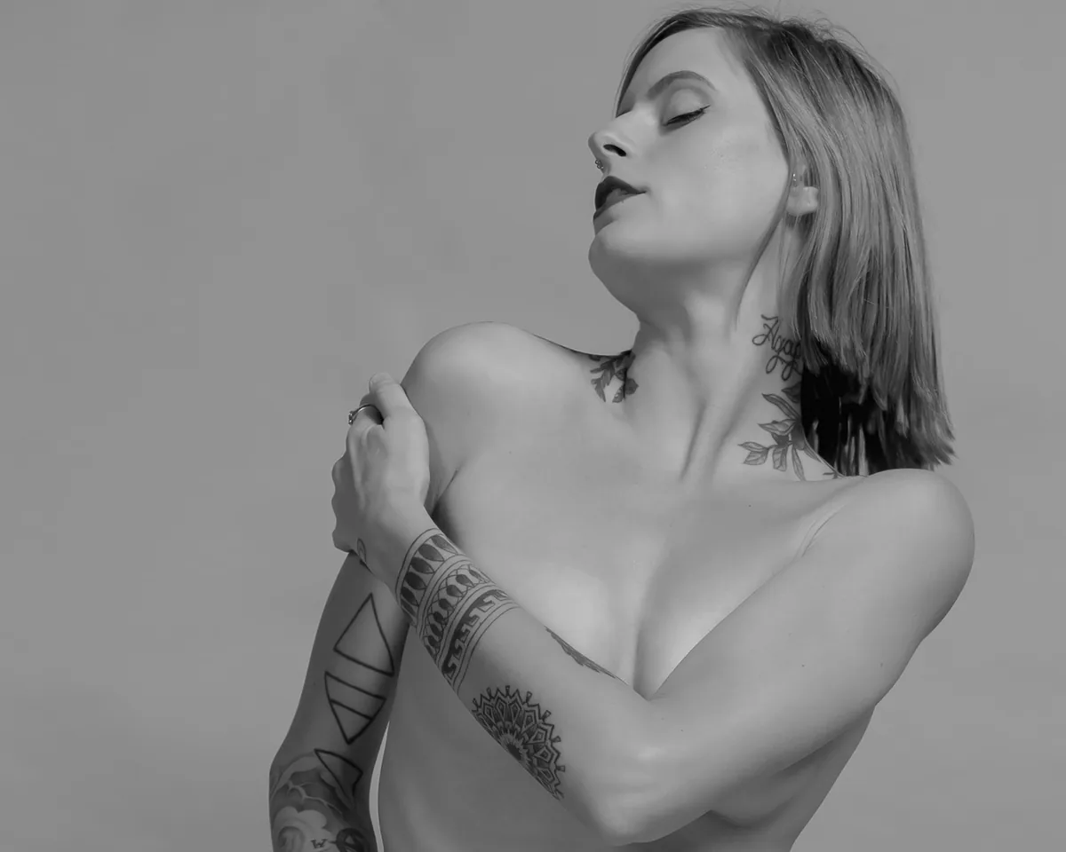 Woman with short hair and various tattoos on her neck and arm, posing with eyes closed and head slightly tilted back, one arm crossed over her chest.