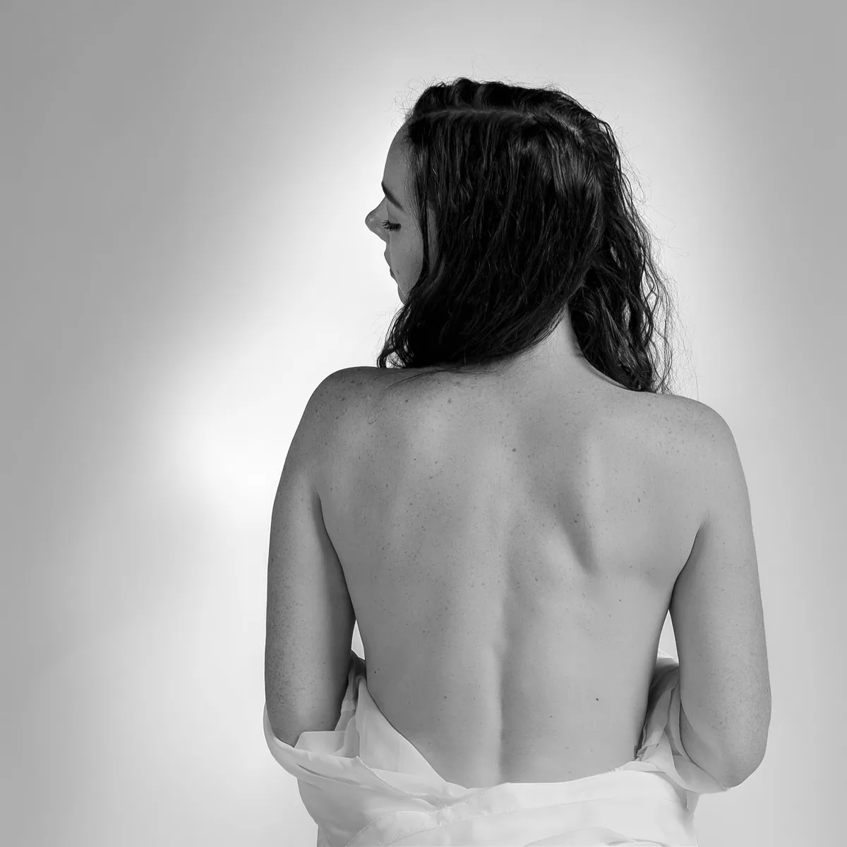 Back of a woman with long dark hair looking to the side, bare shoulders visible against a light gray background, wearing a white garment loosely draped around her lower back