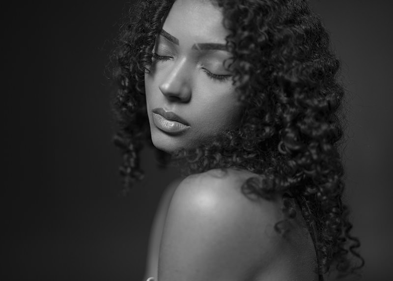 Close-up of a woman with curly hair and closed eyes, facing sideways, shot in black and white with soft lighting highlighting her shoulder and face.