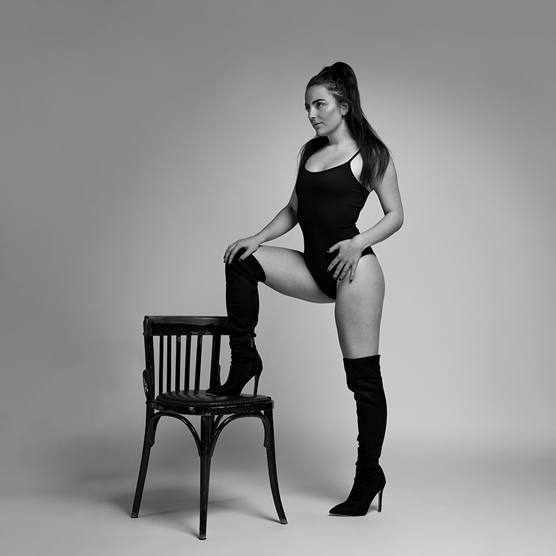 Woman standing with one leg raised on a wooden chair, wearing a black bodysuit and thigh-high boots, against a plain background
