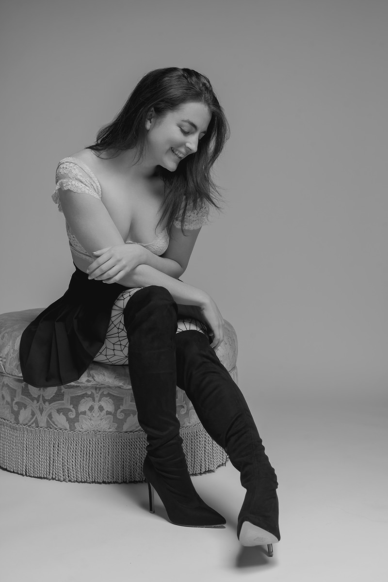 Young woman sitting on an upholstered round stool, wearing a lace short-sleeved top, black skirt, patterned tights, and black thigh-high heeled boots, smiling with crossed arms and looking downward in a studio setting