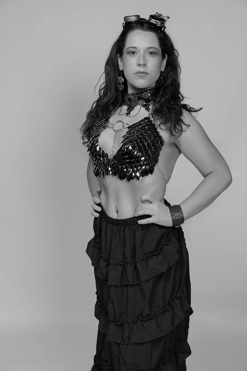Woman with curly hair wearing silver steampunk goggles, a black metal scale bra, a lace choker with gears, dangling earrings, and a ruffled long skirt, posing with hands on hips against a plain background