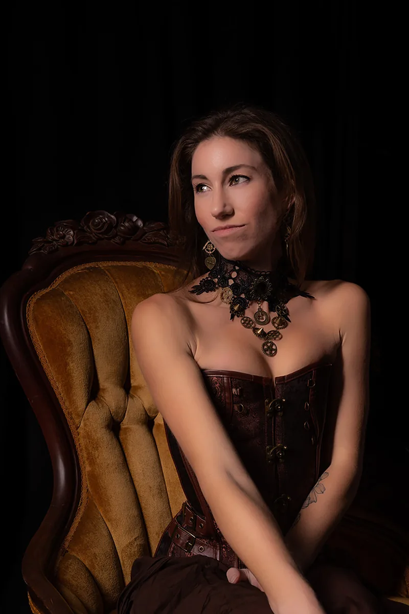 Woman seated on a vintage tan velvet chair wearing a dark brown corset and intricate steampunk necklace, looking thoughtfully to the side against a black background
