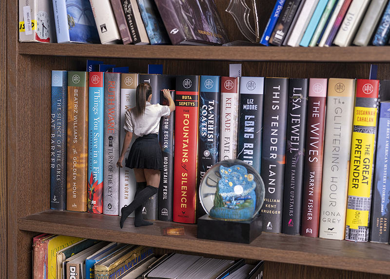 A miniature woman wearing a white blouse and black skirt stands on a wooden shelf, reaching for a large book on a bookshelf filled with oversized books. A decorative snow globe with a blue starry night design is also visible on the shelf.