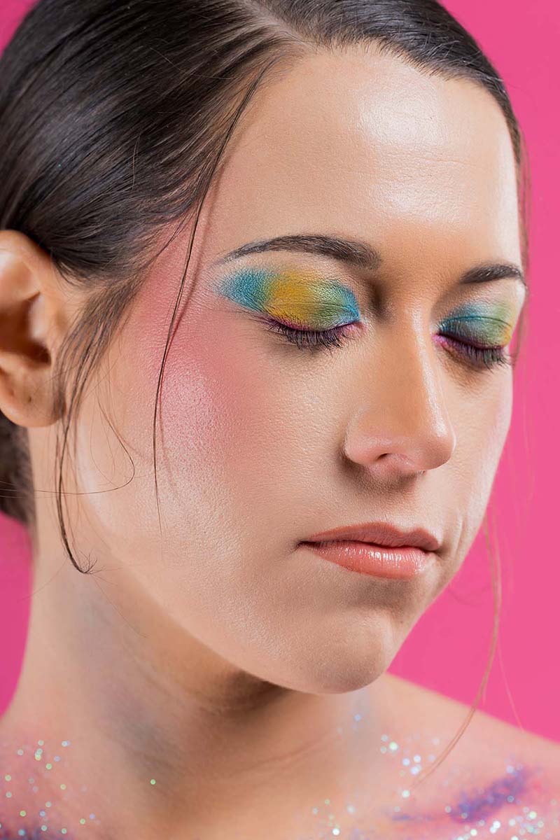 Woman with eyes closed wearing colorful eyeshadow in shades of blue, yellow, and green, light pink blush on cheeks, and subtle makeup on lips, against a pink background.