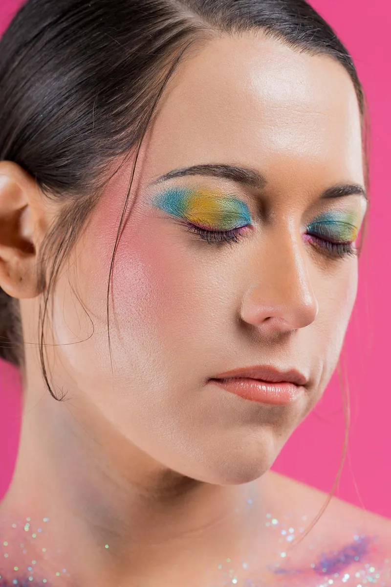 Woman with eyes closed wearing colorful eyeshadow in shades of blue, yellow, and green, light pink blush on cheeks, and subtle makeup on lips, against a pink background.